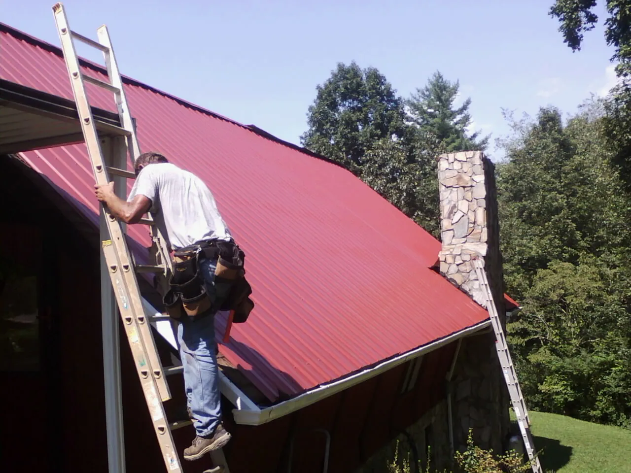 Roofer on a ladder beside a red metal roof and stone chimney — Soffit & Fascia Repair by Roofing Brothers in Corona Del Mar